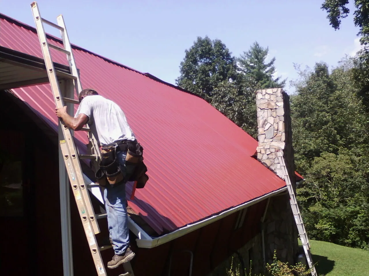 Roofer on a ladder beside a red metal roof and stone chimney — Soffit & Fascia Repair by Roofing Brothers in Burnt Ranch